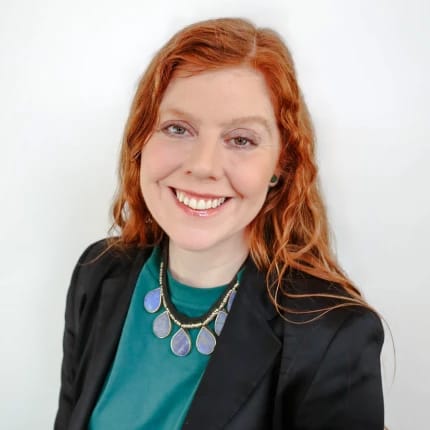 A headshot of a woman with red hair, smiling, wearing a black blazer over a teal shirt with a chunky blue necklace, against a white background