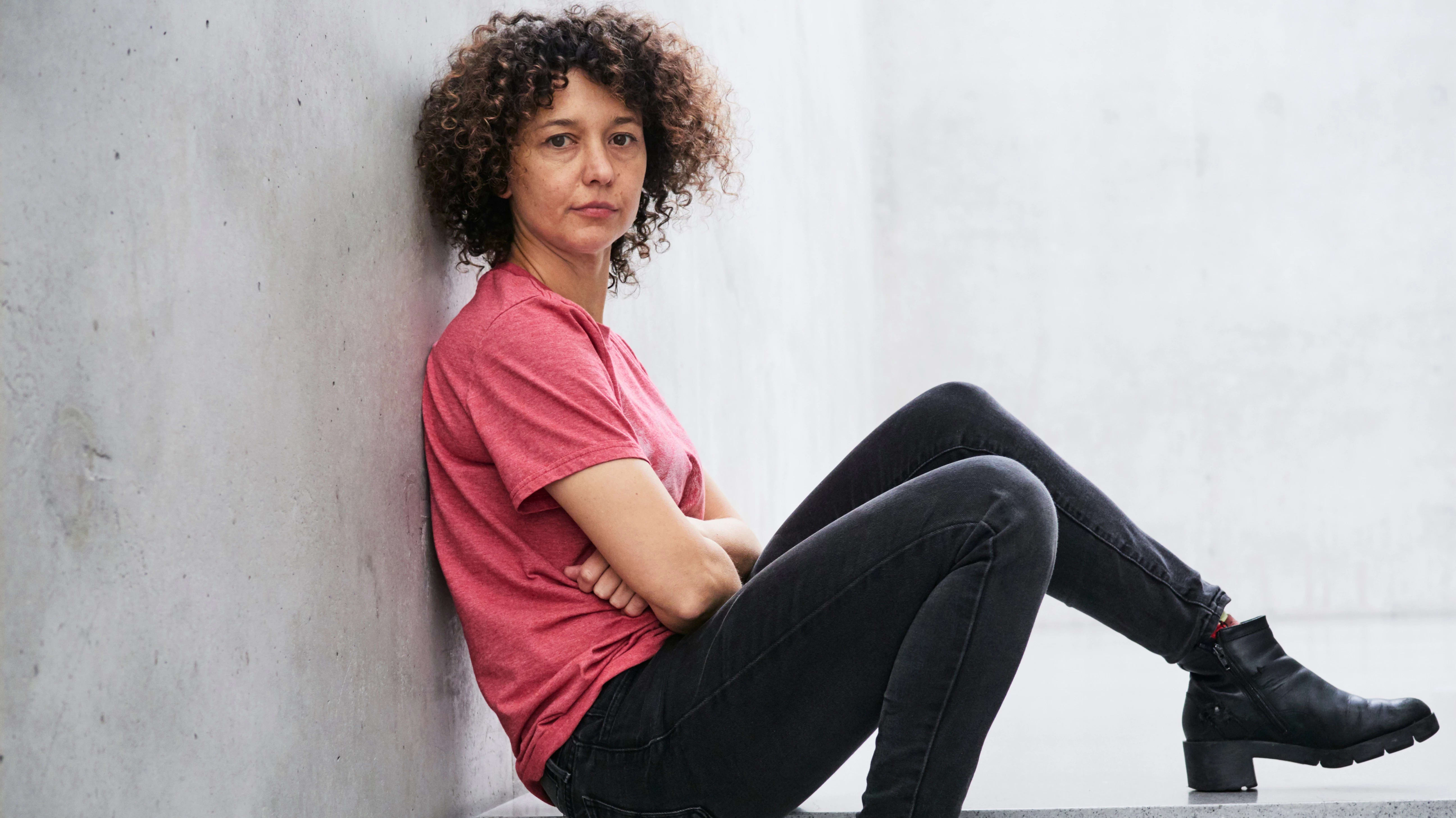 A photograph of Mika Rottenberg. Mika is sitting on a cement step with her back against the wall. She is wearing a red t-shirt with black jeans and boots. Her head is turned to the camera with a relaxed expression.