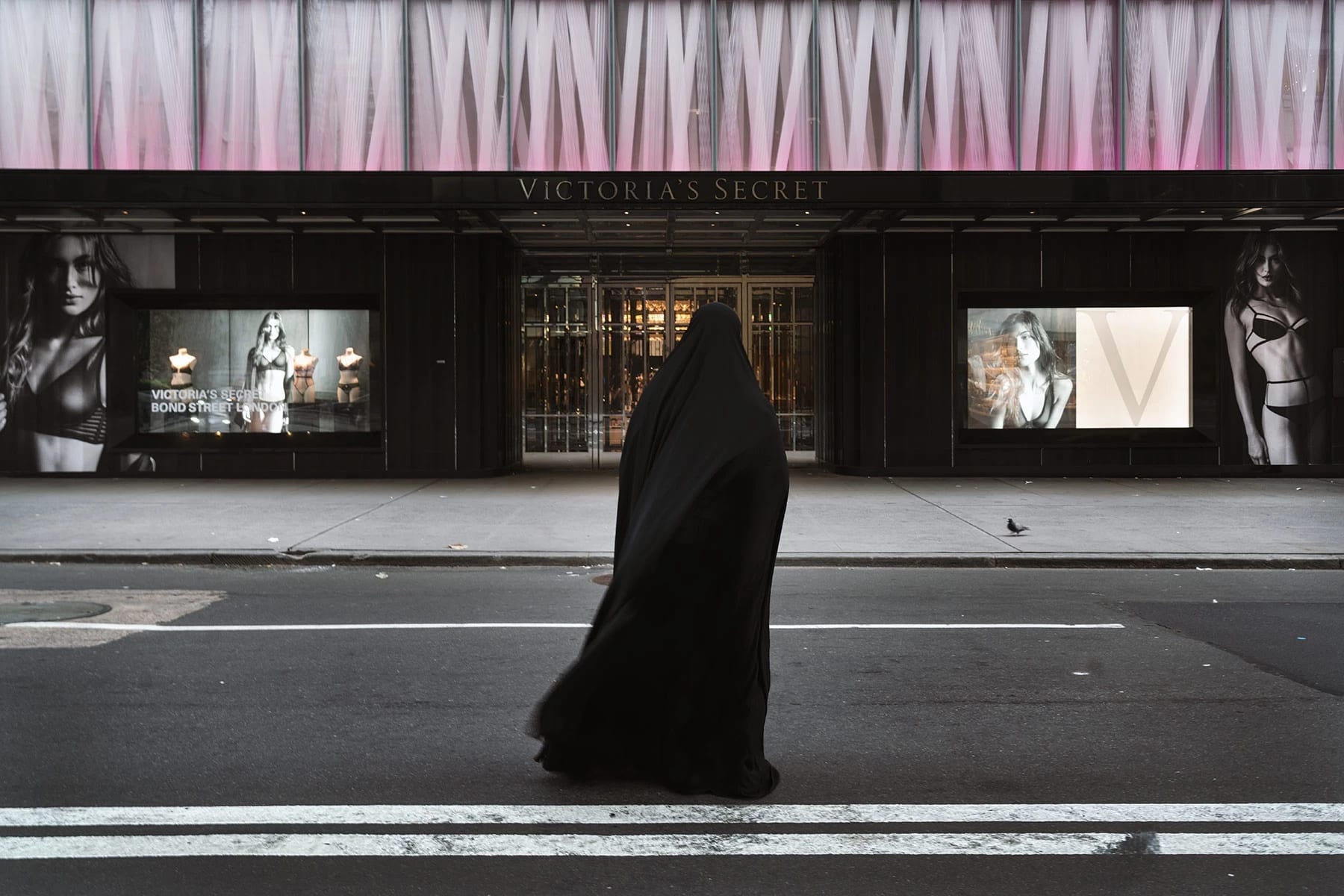 A color photograph of a figure enveloped in a flowing black garment walks across a city street, directly in front of a large Victoria's Secret store. The storefront features large, illuminated advertisements of models in lingerie on either side of its main entrance.