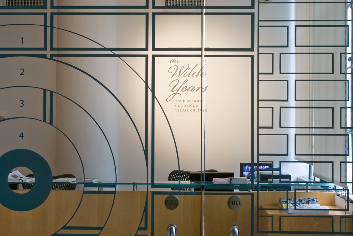 Photograph from outside SVA Chelsea Gallery glass doors, looking inside, with exhibition title visible behind desk.