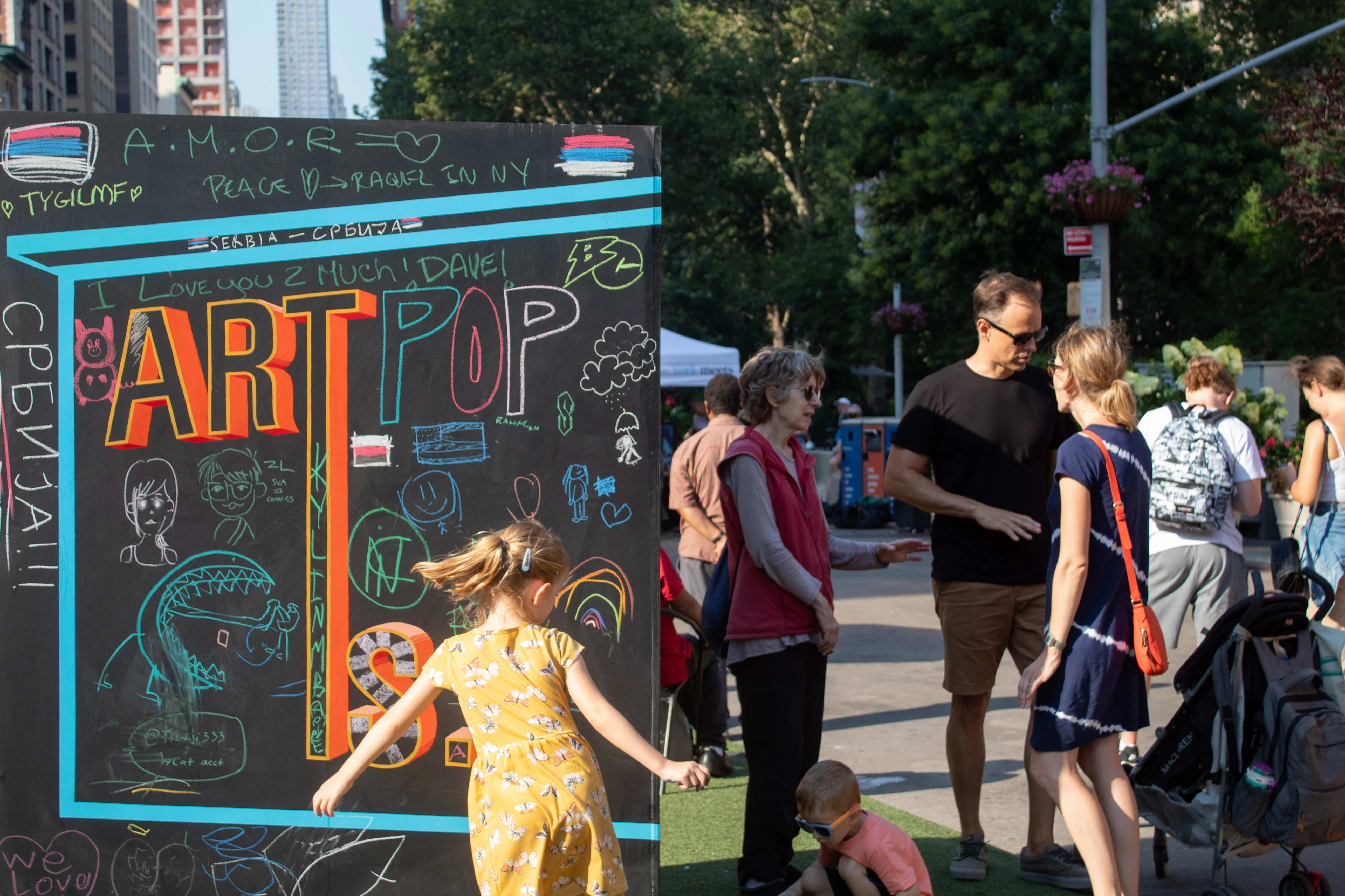 An image of the black chalkboard box with the words "Art Is..." with writings and drawings in chalk from the public. A family enjoying Salsa and Art Nights on the Flatiron North Plaza. 