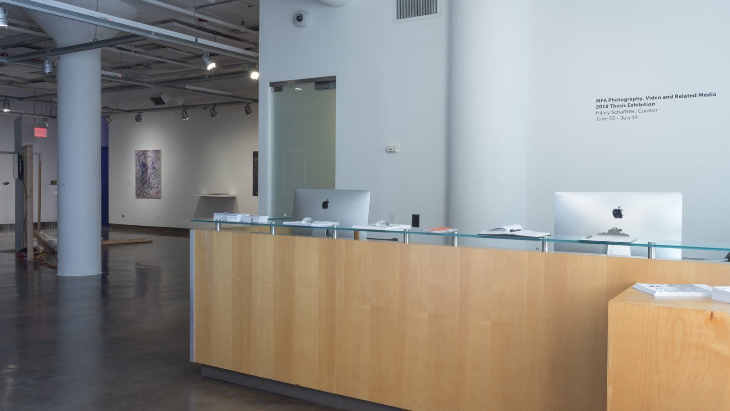 Photo of exhibition installation. View from the gallery entrance, with gallery desk and two computers in the center of the frame and on the far left are glimpses of student artwork.