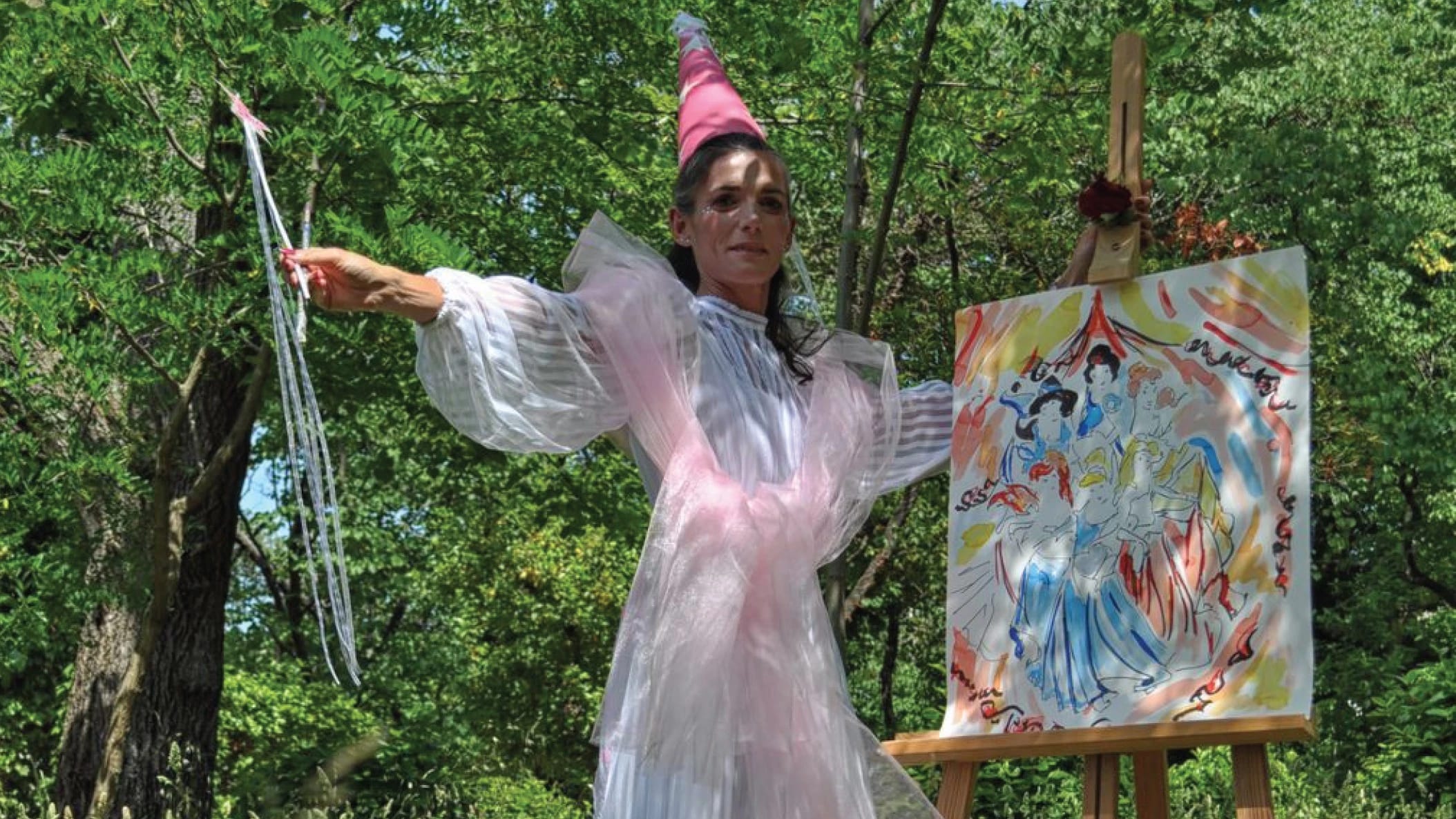 photograph of a woman in a fairy costume standing beside an easel outside.