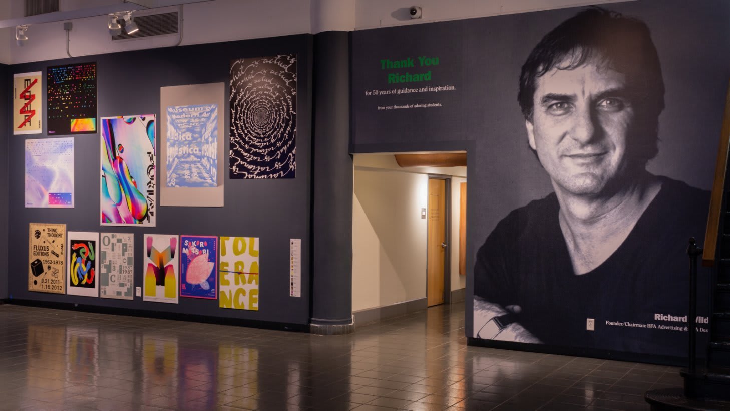 View from entrance of SVA Gramercy Gallery with multiple prints hung "salon"-style to the left, a hallway in the center of the frame, and a wall-sized black and white portrait of Richard Wilde to the right.