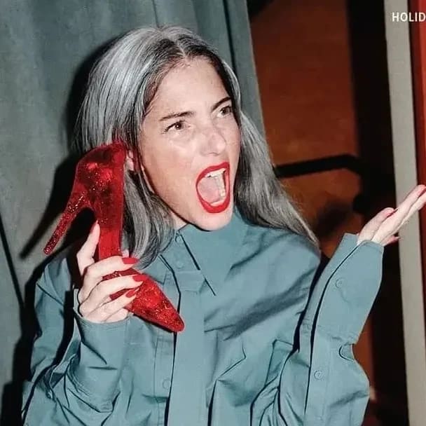 A person sitting backward on a red chair, holding a red high heel to her ear, laughing. Wears a blue shirt and black stockings, with gray hair and red lipstick.