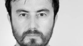 An expressionless, bearded, white man wearing a white button down shirt stairs at the viewer.