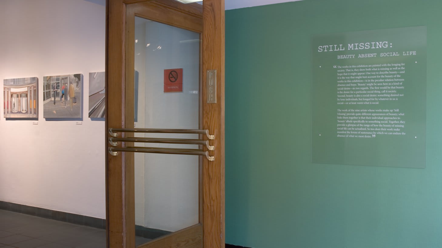 Photo from just inside main doors at Visual Arts Museum, showing the exhibition title and curator’s statement on wall to the left, then just inside the wood and glass entry doors are three paintings.