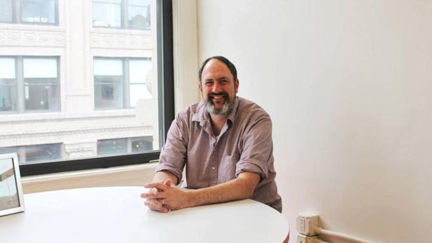 A man sitting in a room with a window in it at a white table smiling with his forearms and hands placed on top of the table.