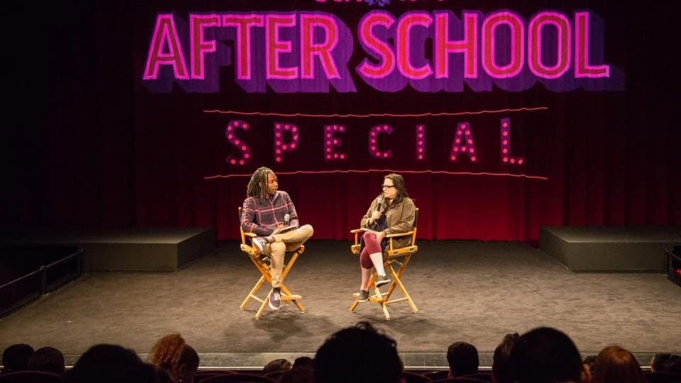 Two people sit on a stage in directors chairs in front of a curtain with the words "After School Special" in pink behind them. There's a crowd facing the stage in the foreground.