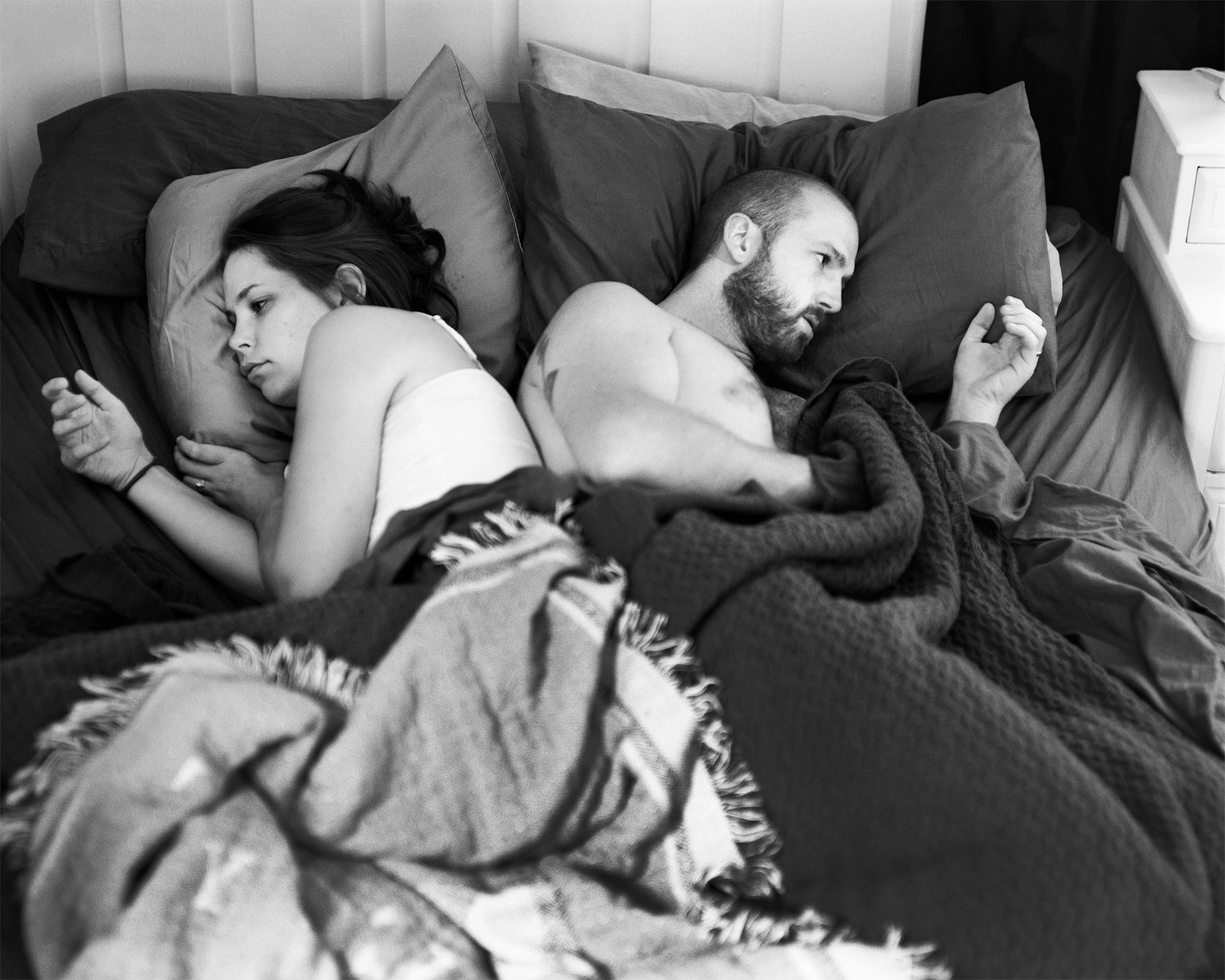 A black and white photograph of a man and a woman lying side by side in bed, facing away from each other. Each holds a hand in the air as if scrolling or holding a phone, though no devices are present. Their expressions are distant and disconnected. The photo is black and white and emotionally charged.