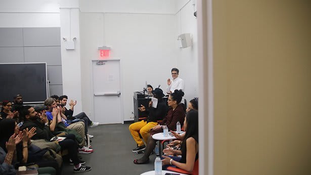 Various people sit in a room facing a row of people. The crowd is applauding.