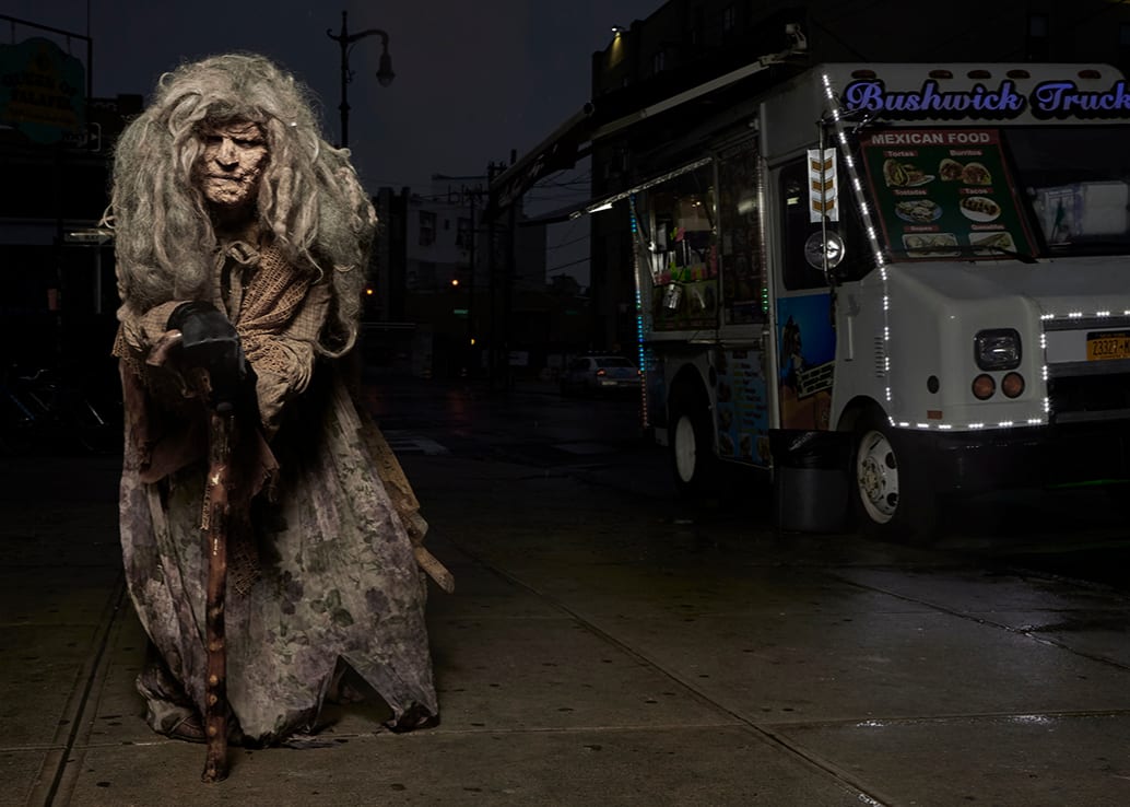 Photograph of the alien Claywoman, hunched over and standing next to taco truck