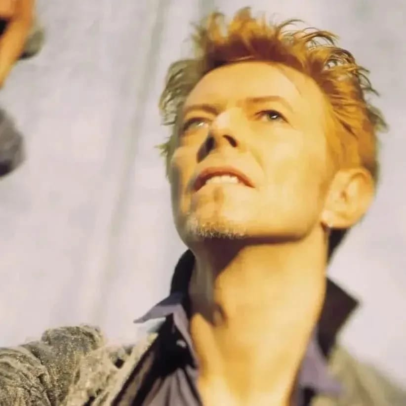 A person with styled light hair and a goatee wears a velvet jacket and shirt, looking upward while holding a microphone stand against a light background.