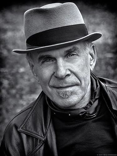 A black and white photo portrait of the photographer Douglas Dubler. He has light skin, is wearing a fedora & a leather jacket over a black top with a silk scarf around his neck.