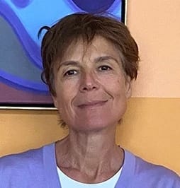 a person with short brown hair and brown eyes smiles in front of a beige wall 