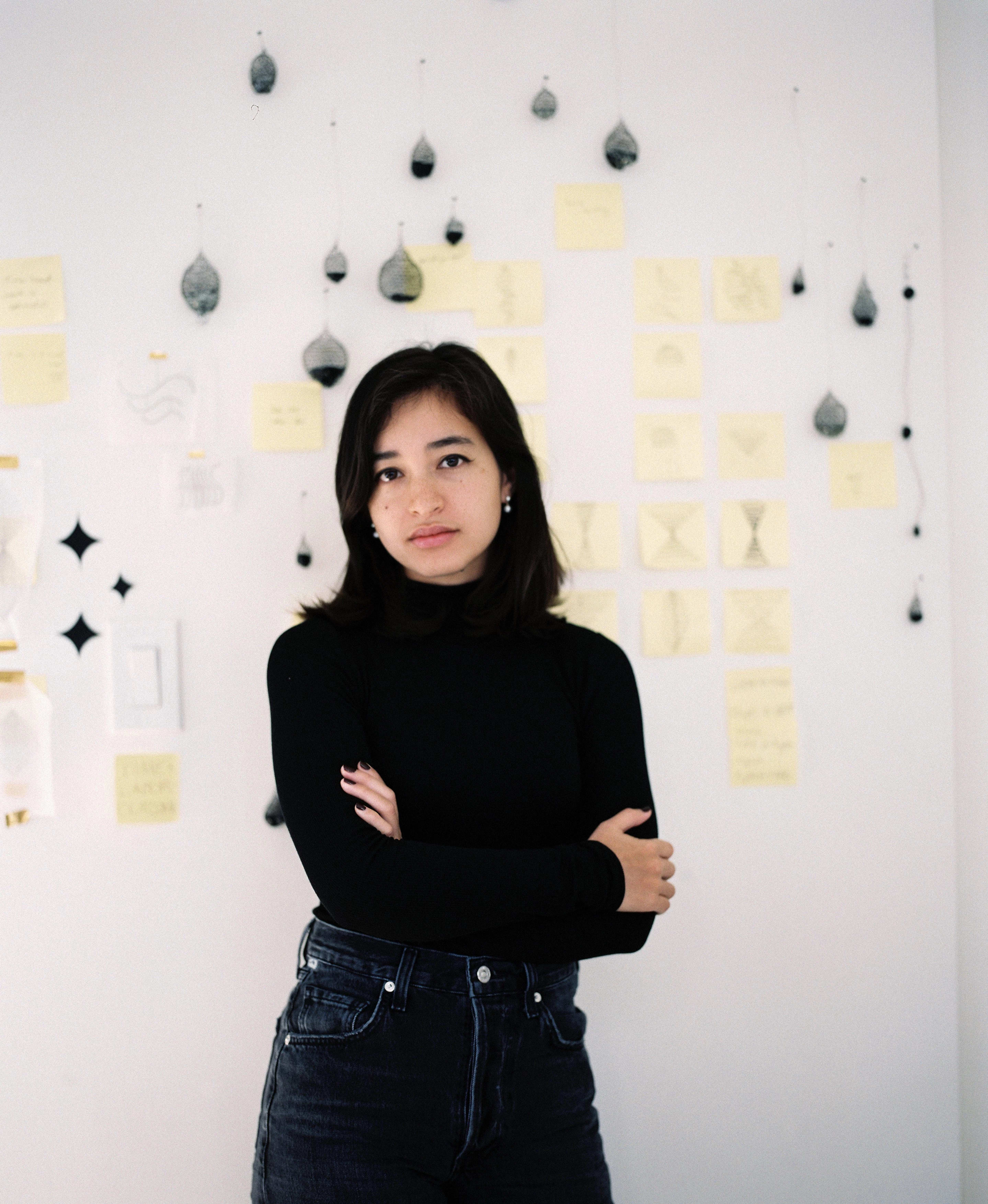 a person with short black hair and a black turtleneck stands with her arms folded