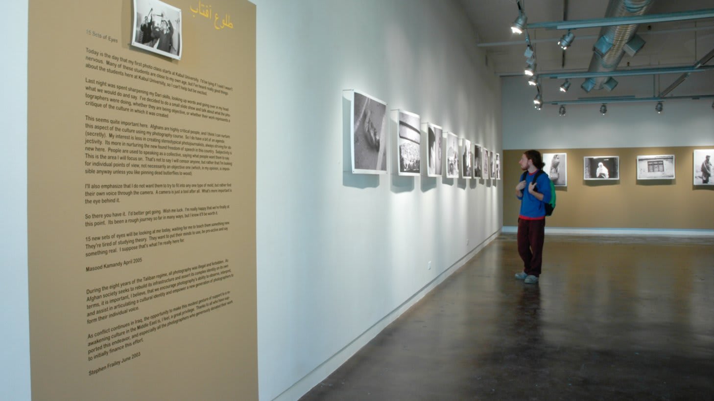 View from back of front room at Visual Arts Gallery, showing seven black and white prints on the left wall, on the right wall (painted gold) are seven additional prints. In the center of the room is a bench.