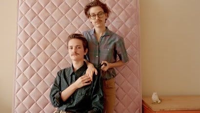 Two brothers both lean up against the mattress in a bedroom.