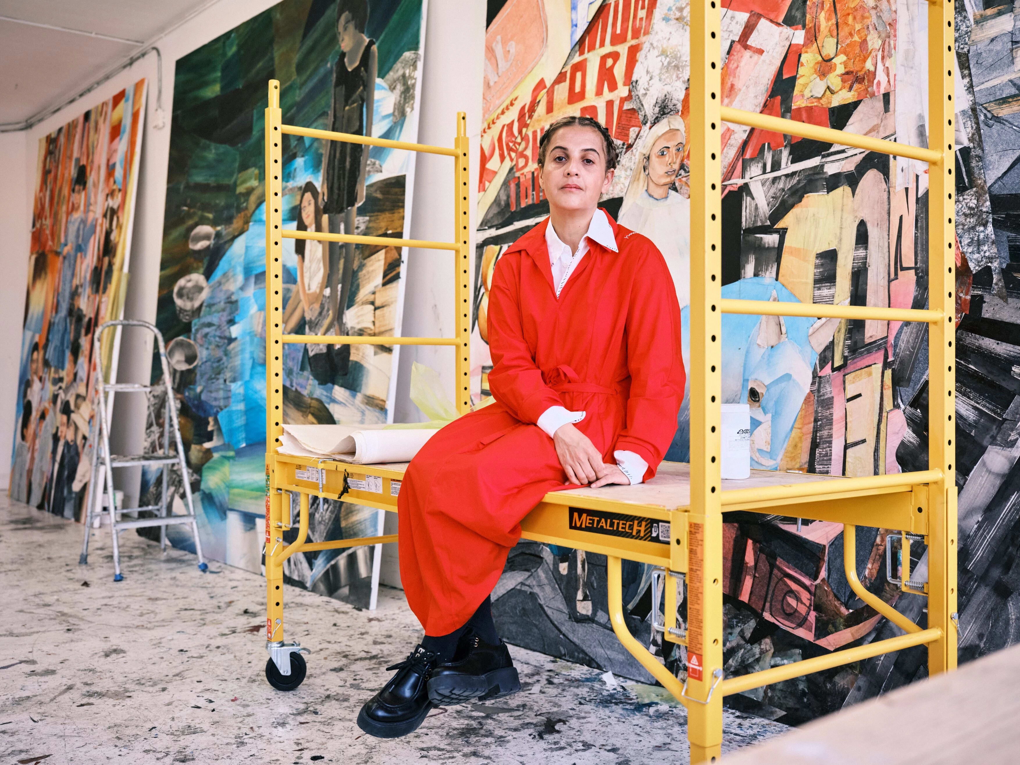 A woman in a bright red jumpsuit sits on a yellow metal frame on wheels in a studio.