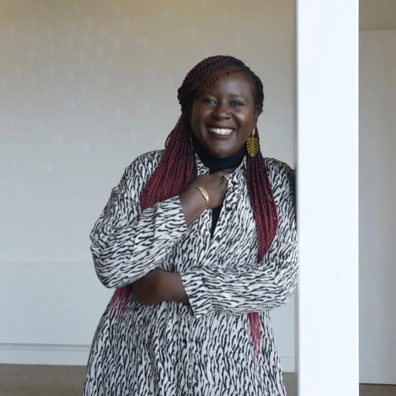 A photo of Fatima Bintou Rassoul SY leaning against a white pillar. She is in a black and white patterned dress with her hand under her chin. 