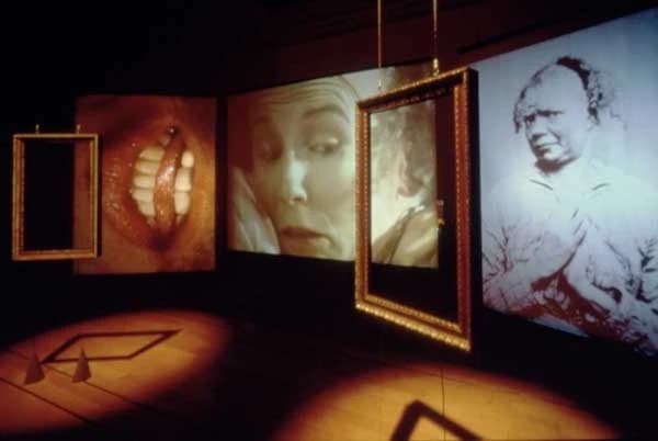 An image of a dark room looking at a diagonal angled wall with several spot lit frames hanging. The frames contain close up portraits. 