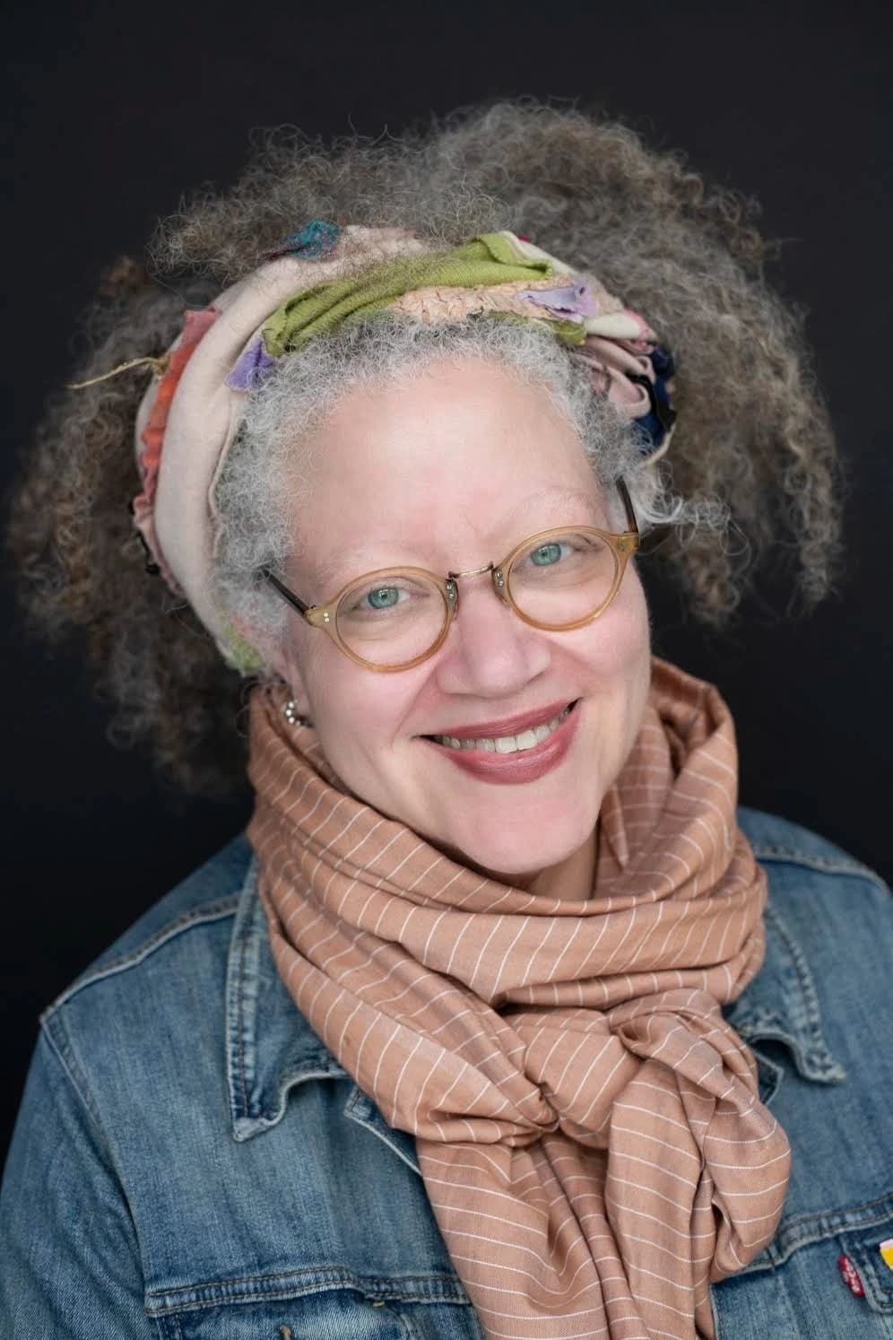 A woman with a bandana in her gray hair smiles, wearing a jean jacket and a colorful scarf.