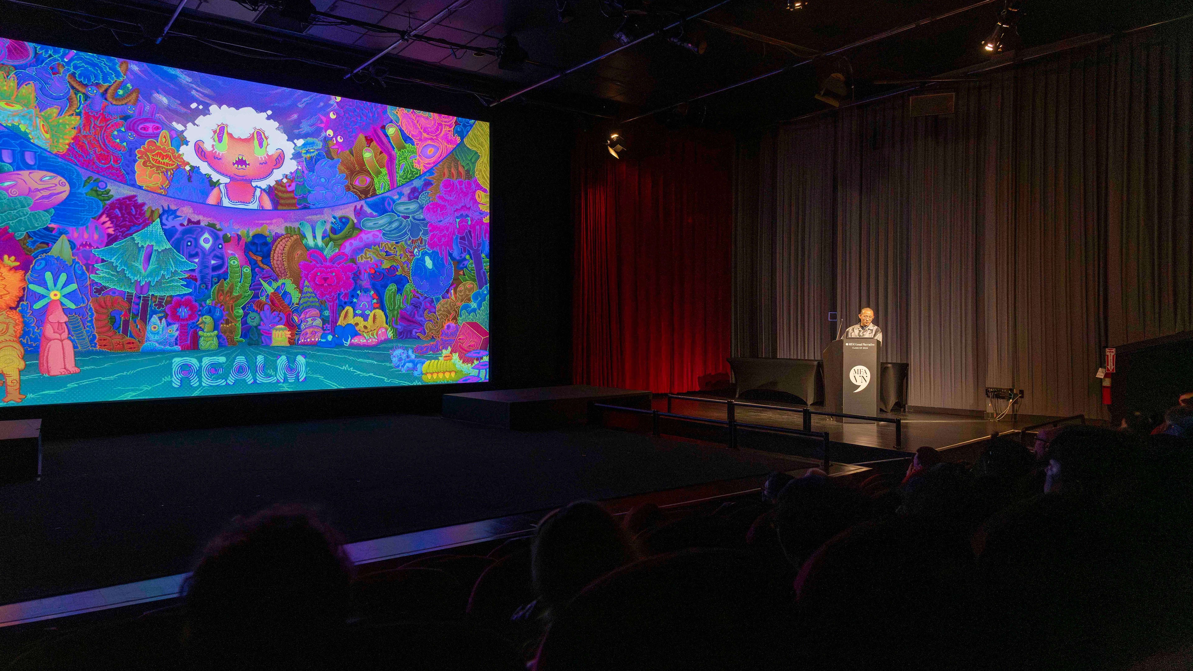 Wide shot of student standing in front of a podium in a large theatre. An illustration in psychedelic colors is projected onto a large screen.