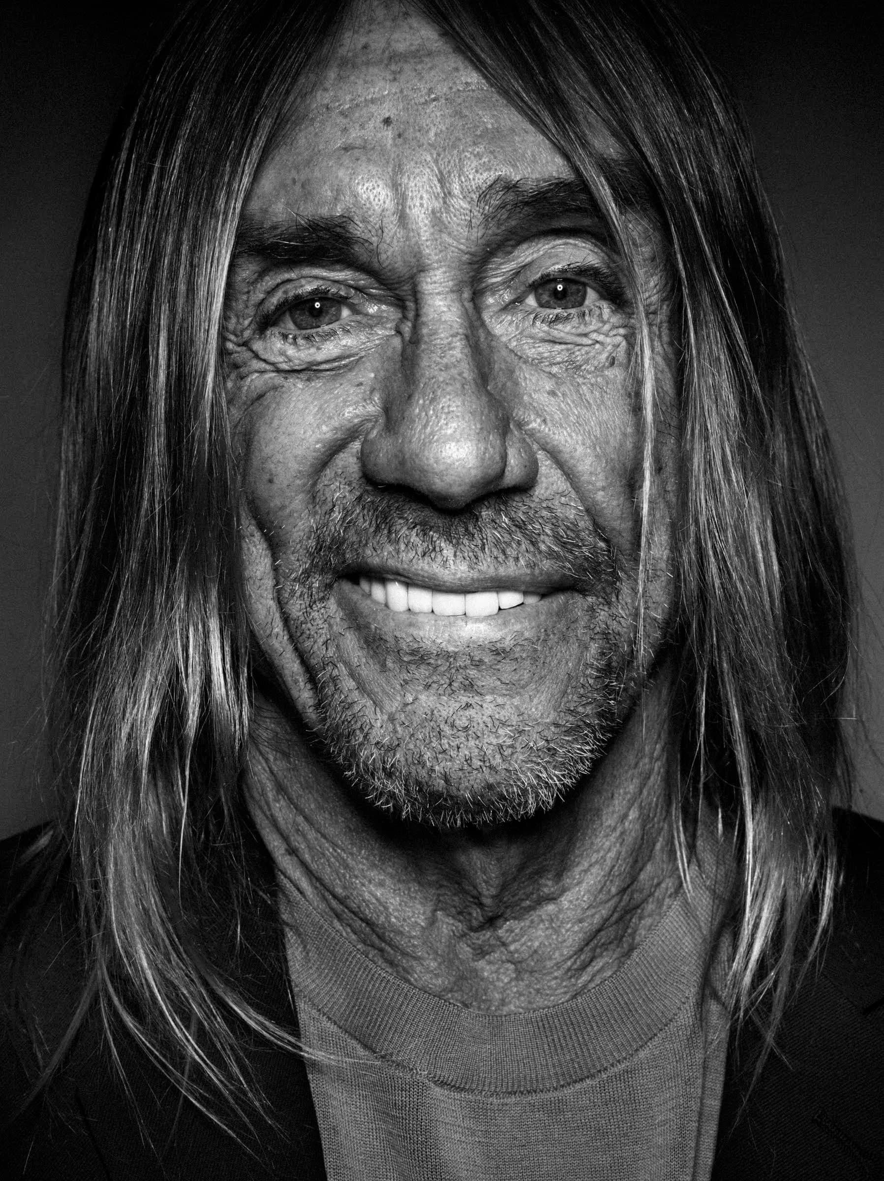 A black and white photo portrait of the singer Iggy Pop, who is looking straight into the camera with a smile. He has long hair and light skin, and is wearing a black cardigan over a gray t-shirt.