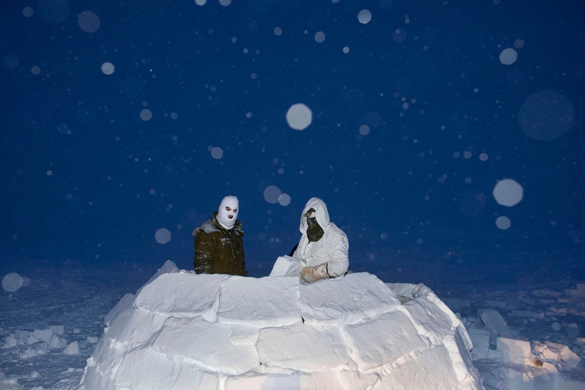 A color photograph of Two soldiers, wearing masks in an igloo made of snow blocks, at dusk while it snows, lit with flash.