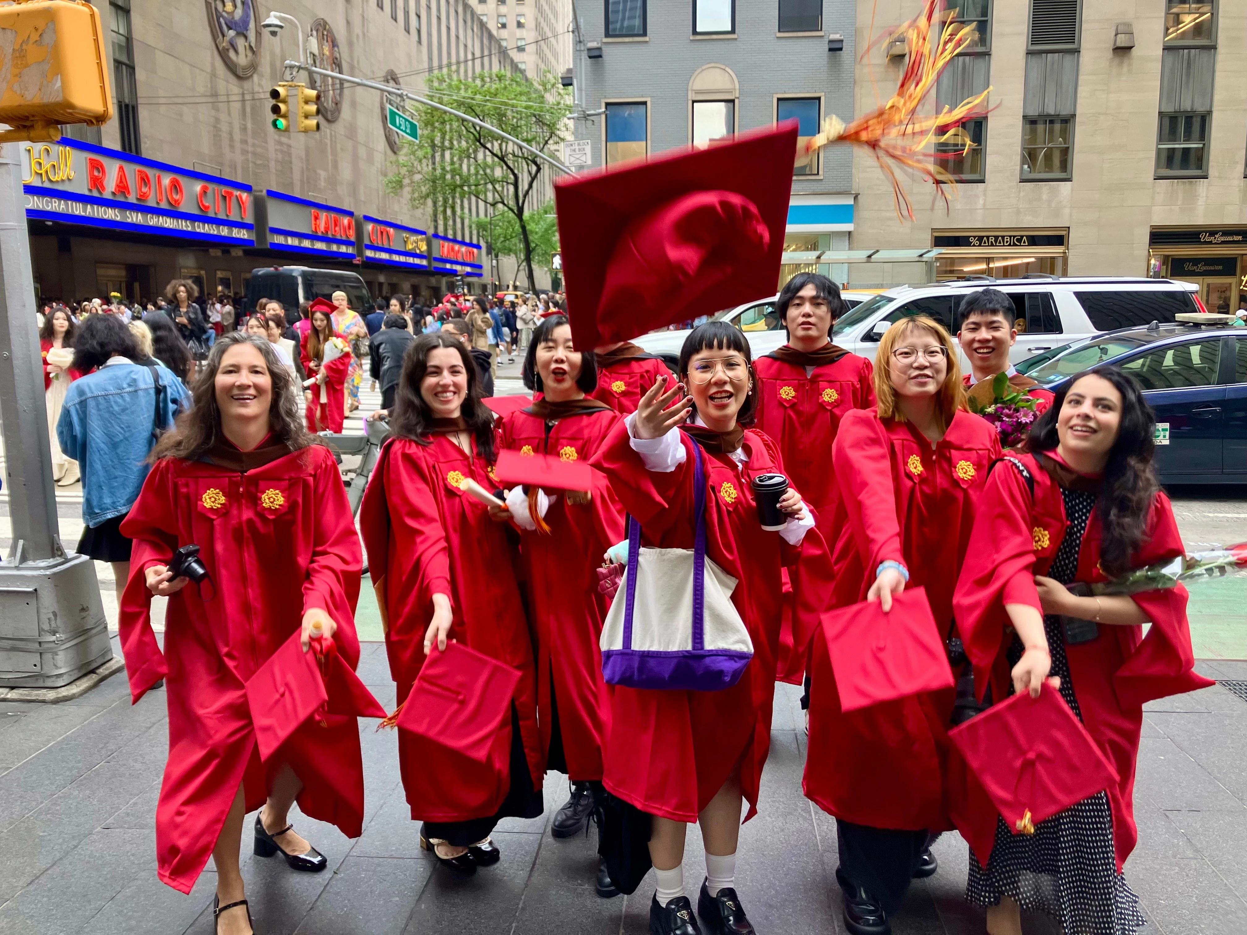 The Masters in Digital Photography graduating class of 2025, in the iconic SVA red graduation gowns, in front of the famed Radio City Music Hall where the commencement ceremony just took place. The students are threwing their caps in excitement. 