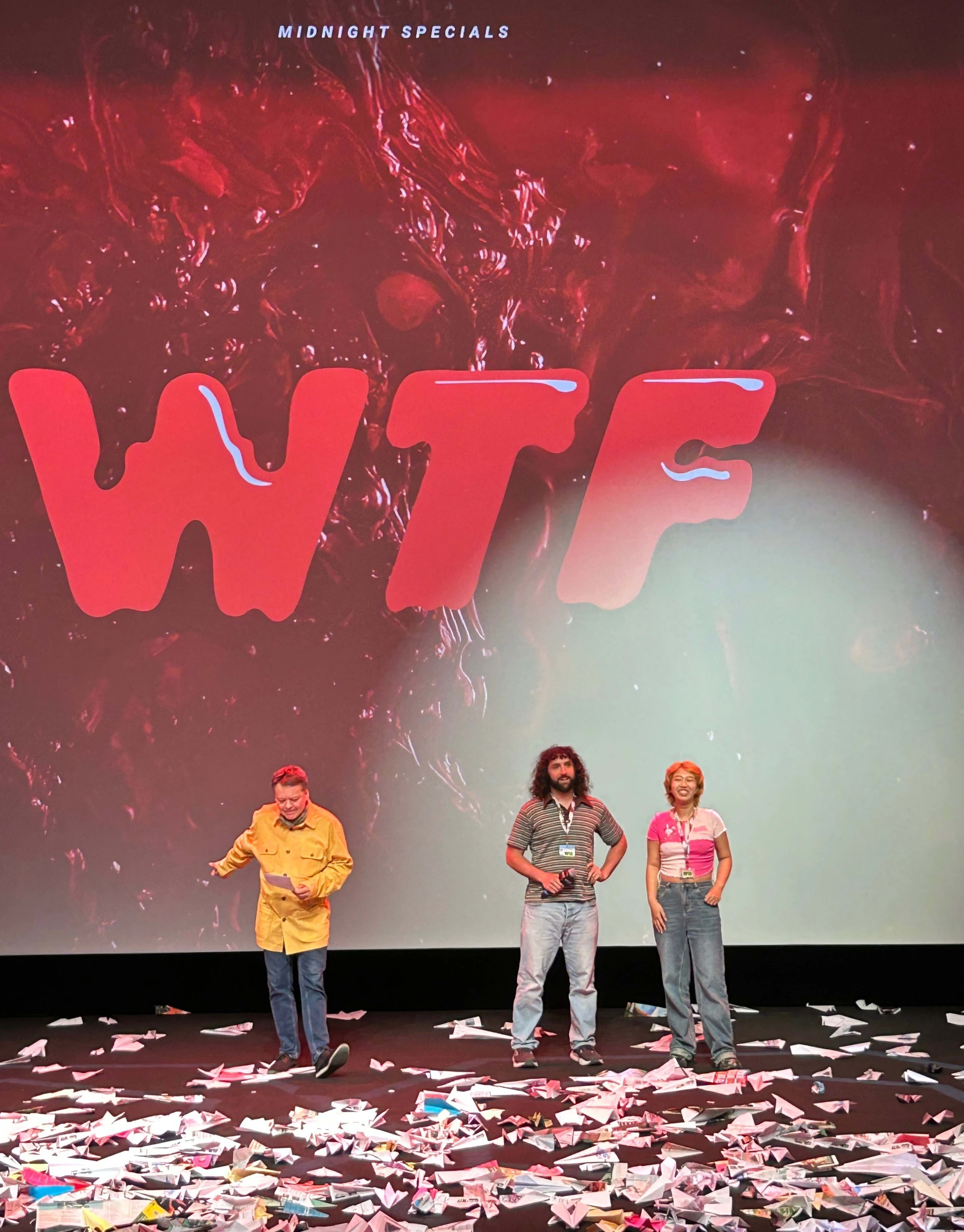 Three people stand on a stage full of paper airplanes with a projection showing WTF behind them