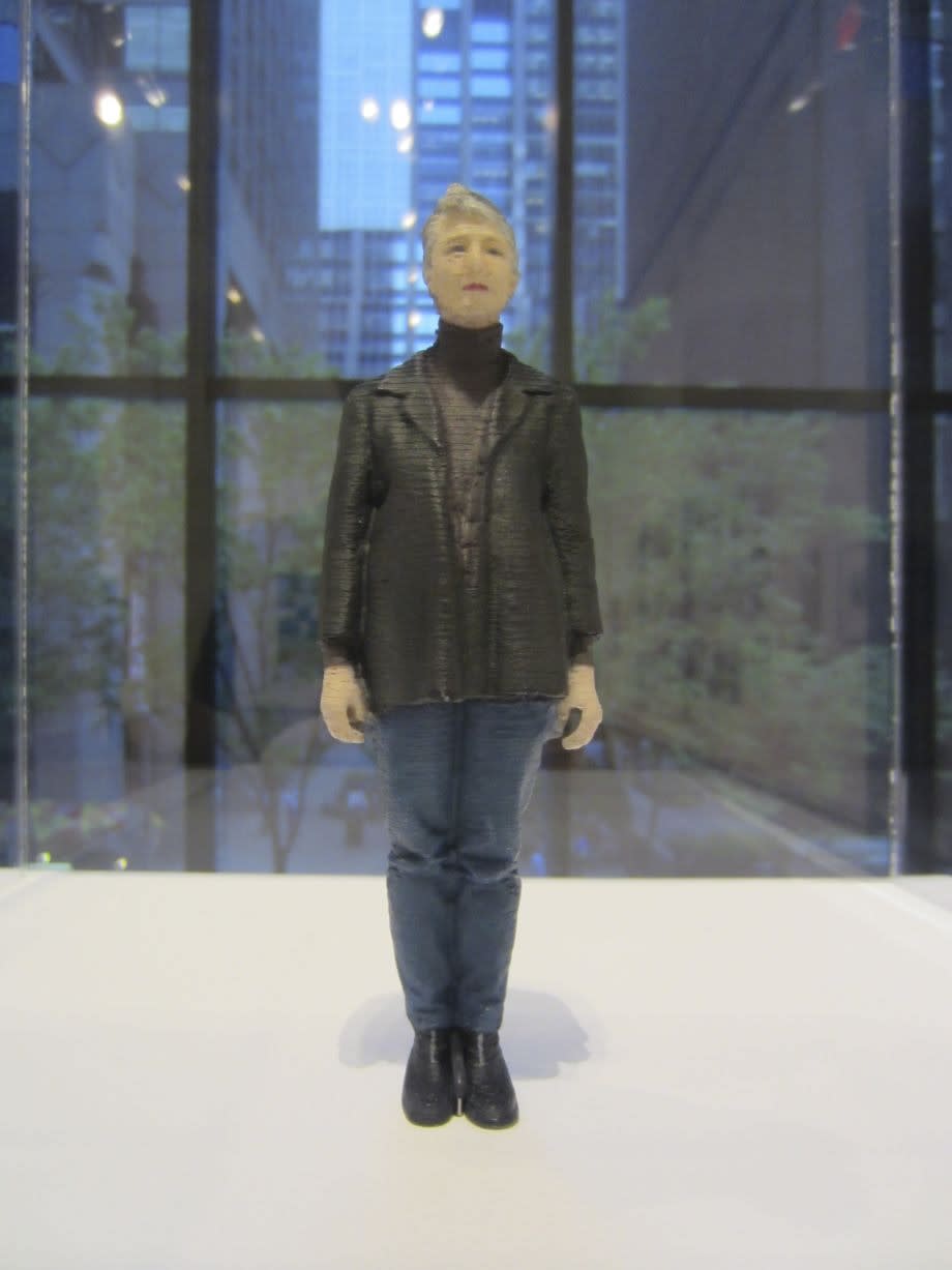 A mini sculpture of Karin Sander in a black suit jacket and jeans that is placed on a white table in front of a window.