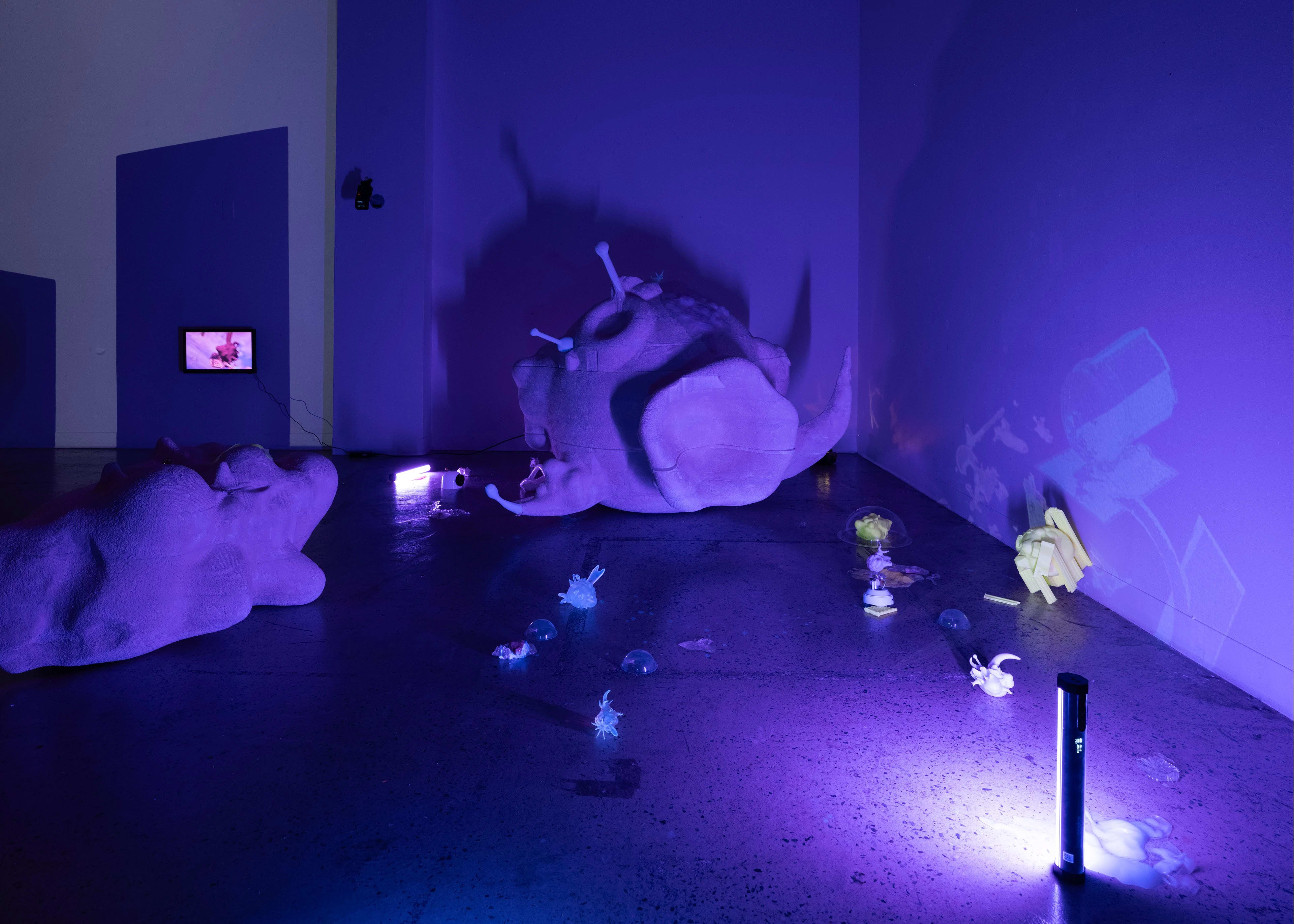 Installation by Kylie Rah. The installation is set in a large room. A large foam sculpture sits in the center of the room and is in the form of a sphere with protruding attachments. There are other small sculptures spread out across the flor. A single dark blue-violet light sits in the corner shining on the objects. The overhead lights are off.