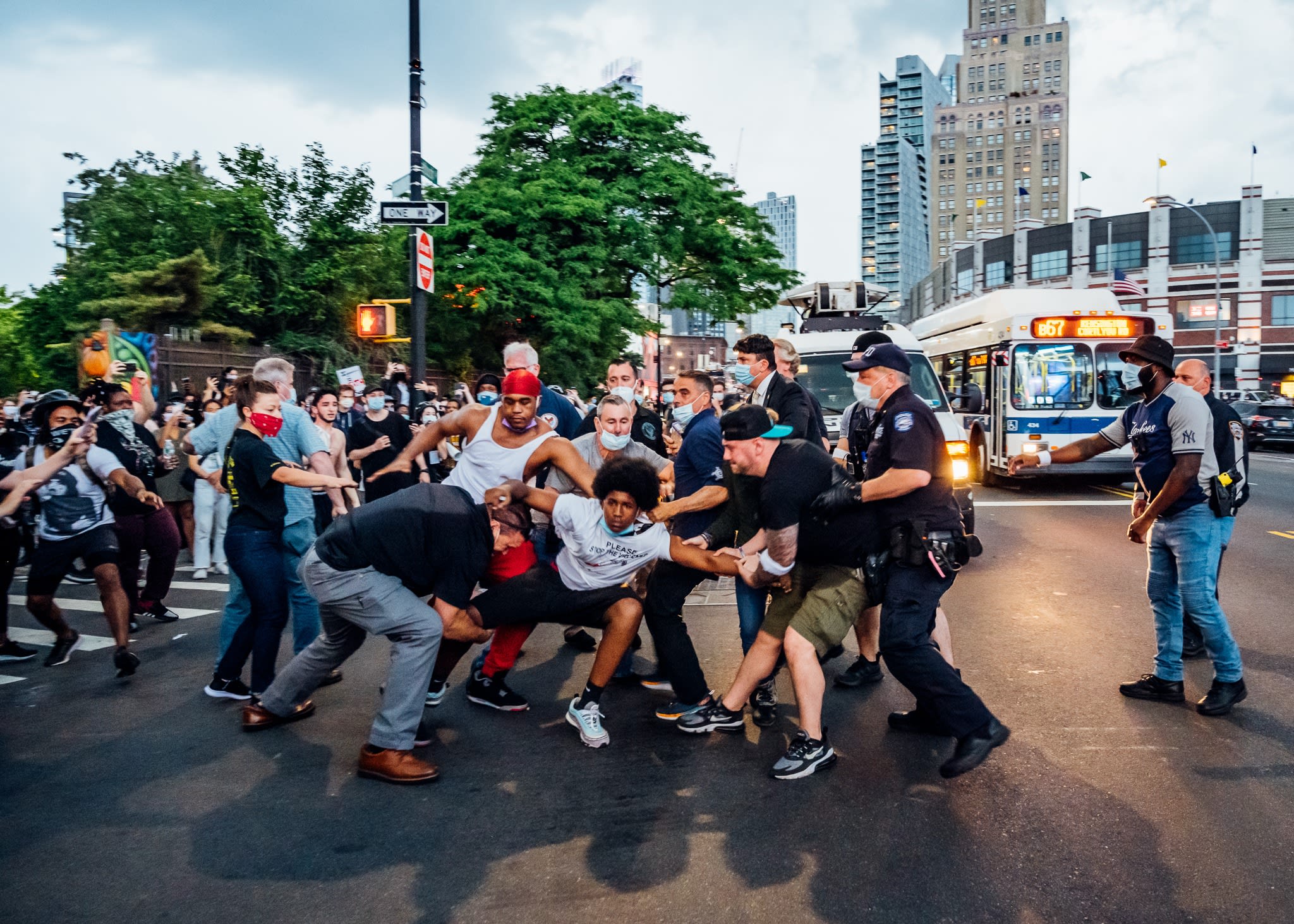 This photo taken of a Black Lives Matter protest is in the middle of a city street, with a confrontation between civilians and police officers. The main subject is an African-American man being tugged by his arm and leg on all sides.