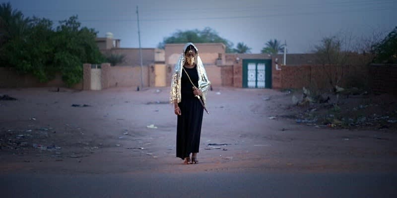 A female presenting person walks away from a gated structure. She wears a black dress that reaches her ankles, a shiny jacket with the hood up, and a gold face covering and she is holding a sheathed knife.