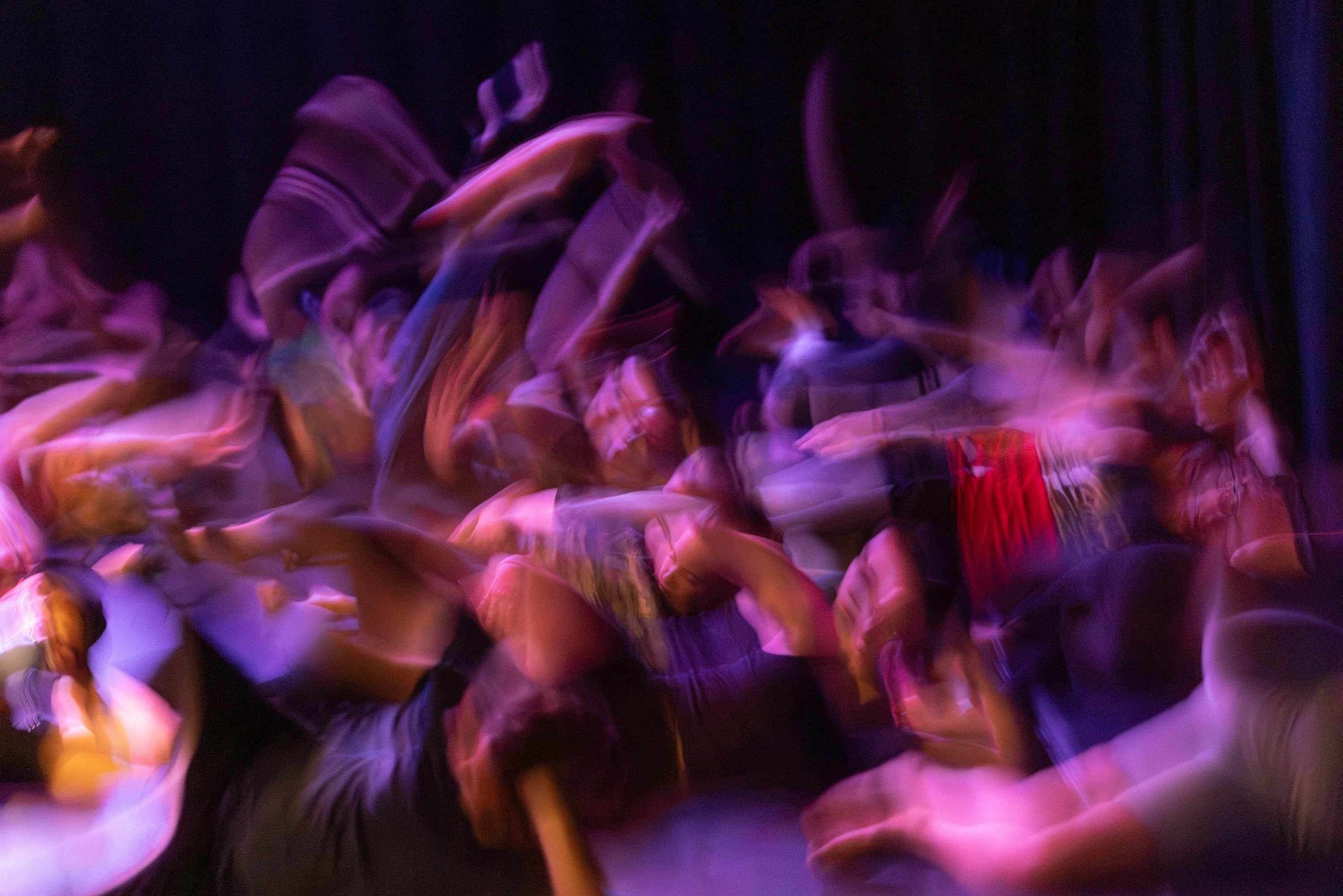 A dynamic, motion-blurred photograph of a crowd in movement under purple and amber stage lighting, suggesting dance or performance.