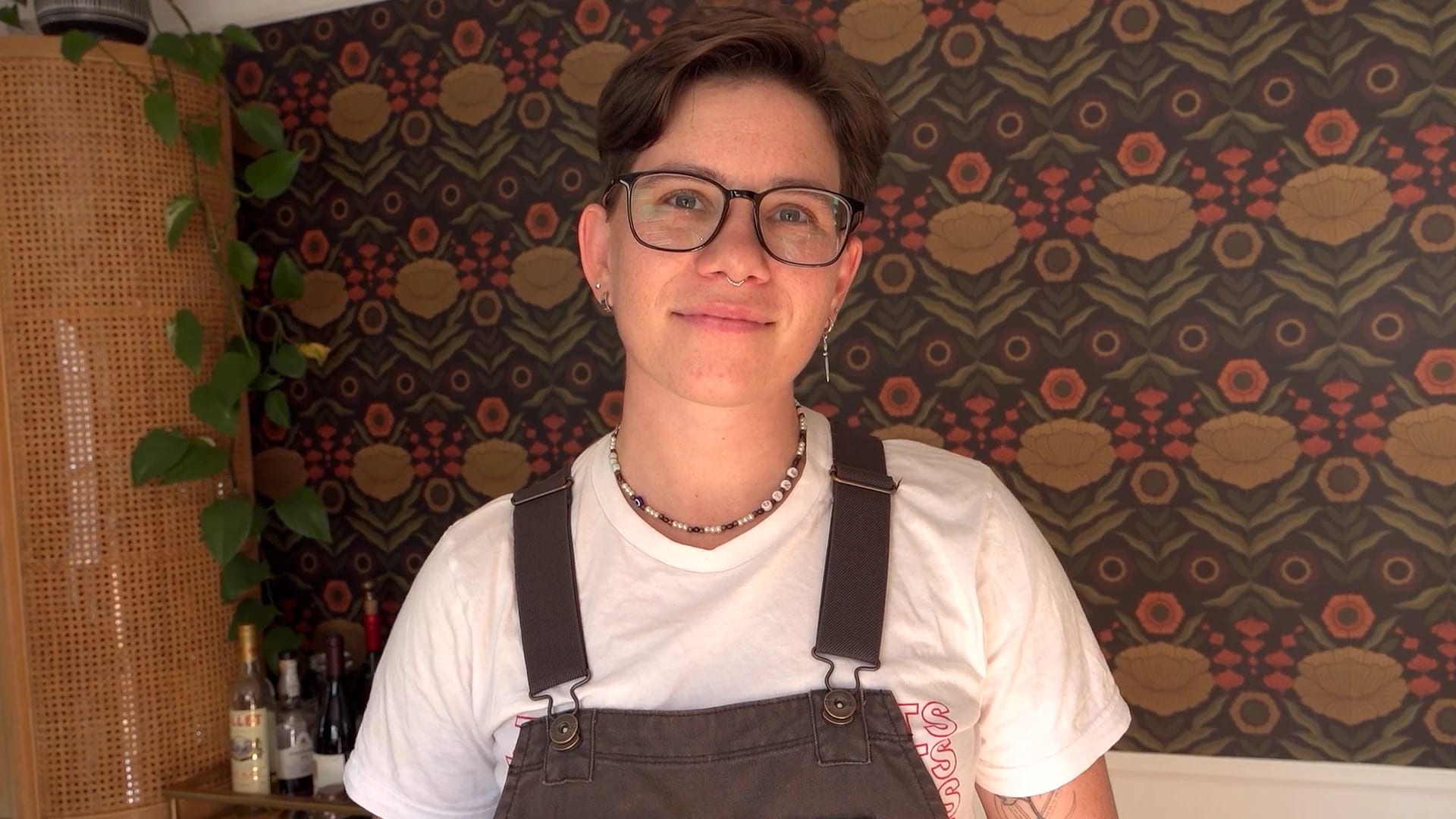 Molly Knox Ostertag smiles at the camera. They're wearing a white tee-shirt and brown overalls. There's a patterned wall behind them and a table with bottles and a plant.