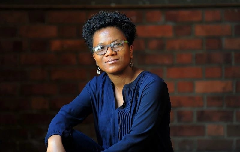 a person in a blue sweater sits in front of a brick wall and smiles at the camera 