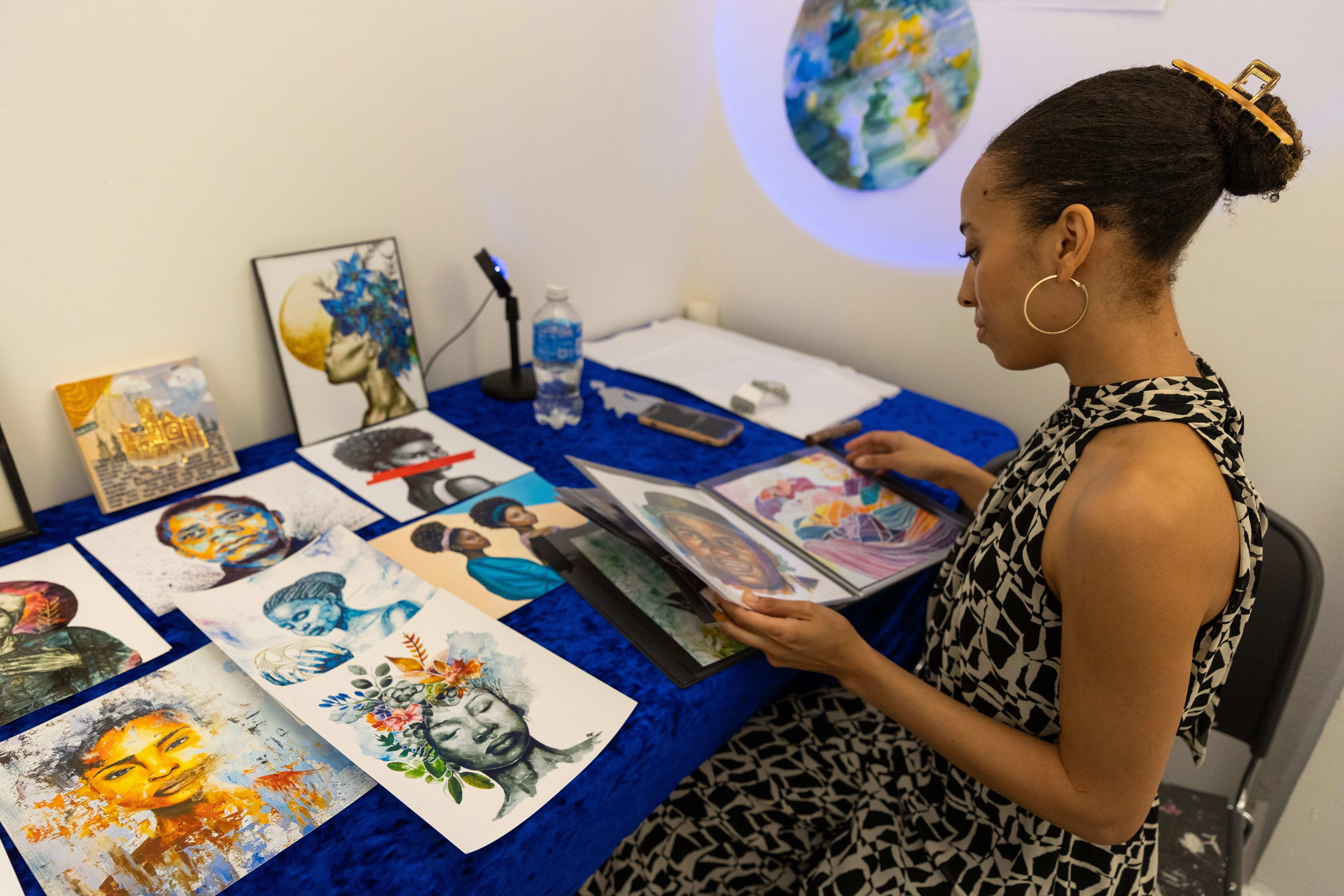 A woman sits at her studio table looking at her various watercolor paintings wearing a geometric black and white dress.