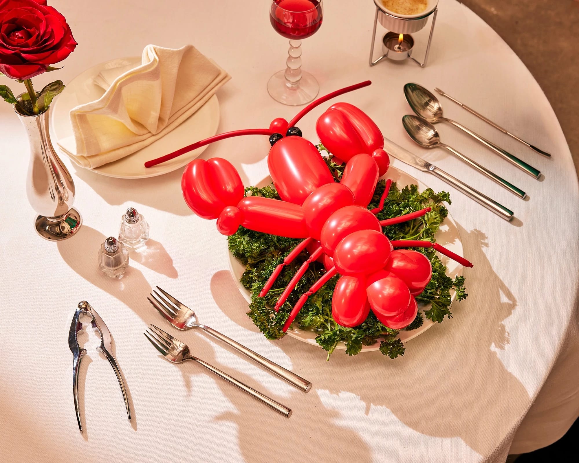 A photograph of a lobster shaped red balloon sitting on top of a bed of kale leaves on a dinner plate. Surrounding the scene is a dinner table fully set up with cutlery,  a dinner napkin folded into a fan, and a single rose in a tin vase.