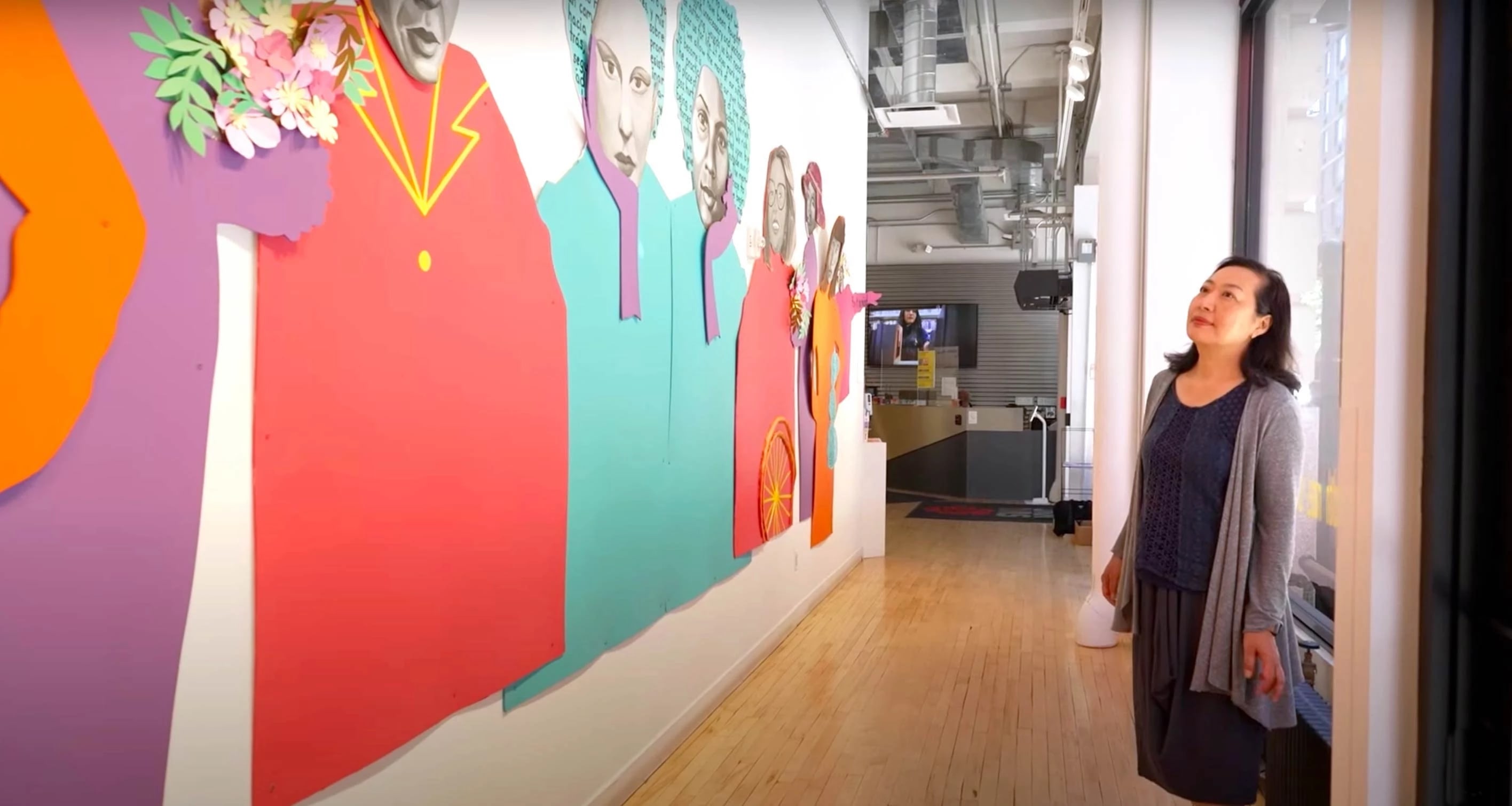 A woman looks up at a large cut-paper collage of several women that has been installed on an interior wall in an SVA campus building.
