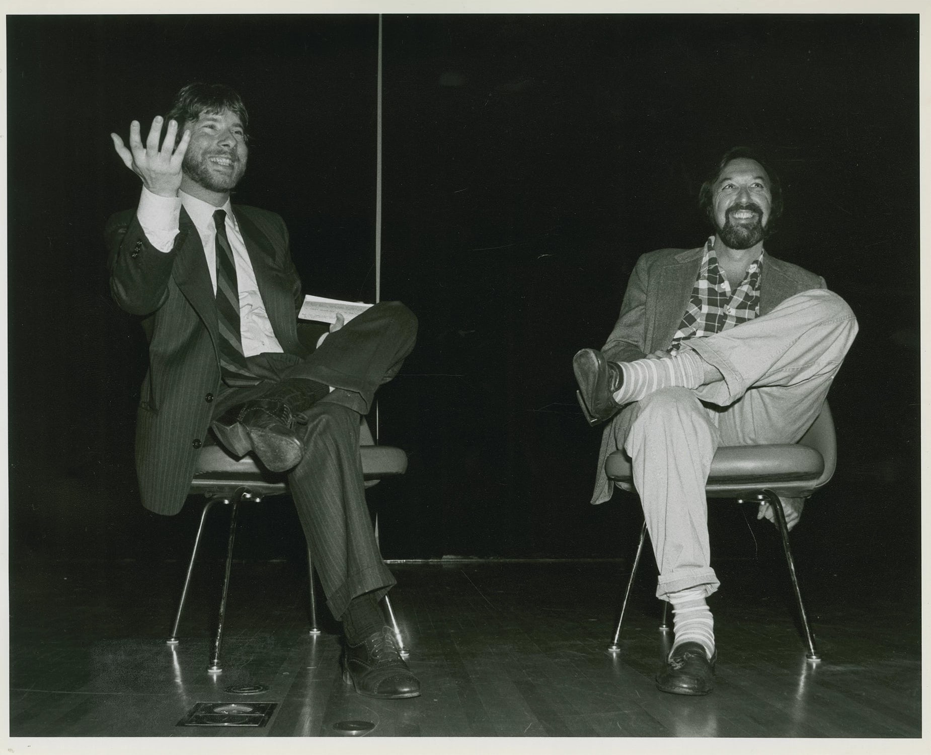 A black-and-white photo of two people sitting on chairs and smiling at something off-camera.