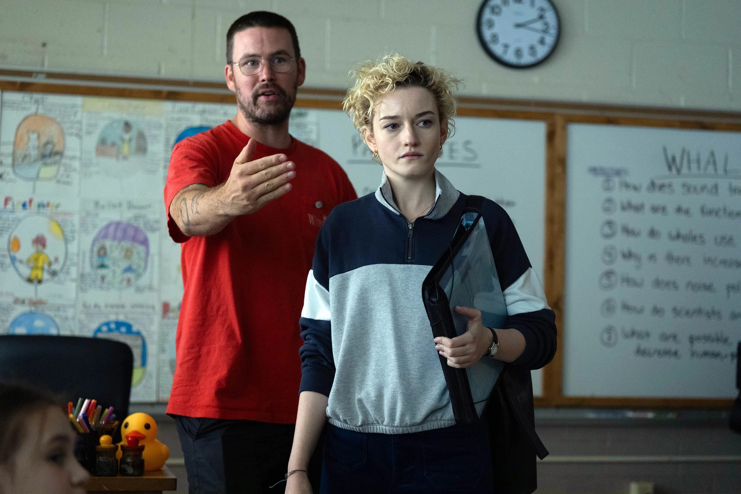 A person in a red shirt gestures in a classroom, appearing explanatory, while another with short curly hair listens intently. A whiteboard and clock are in the background.