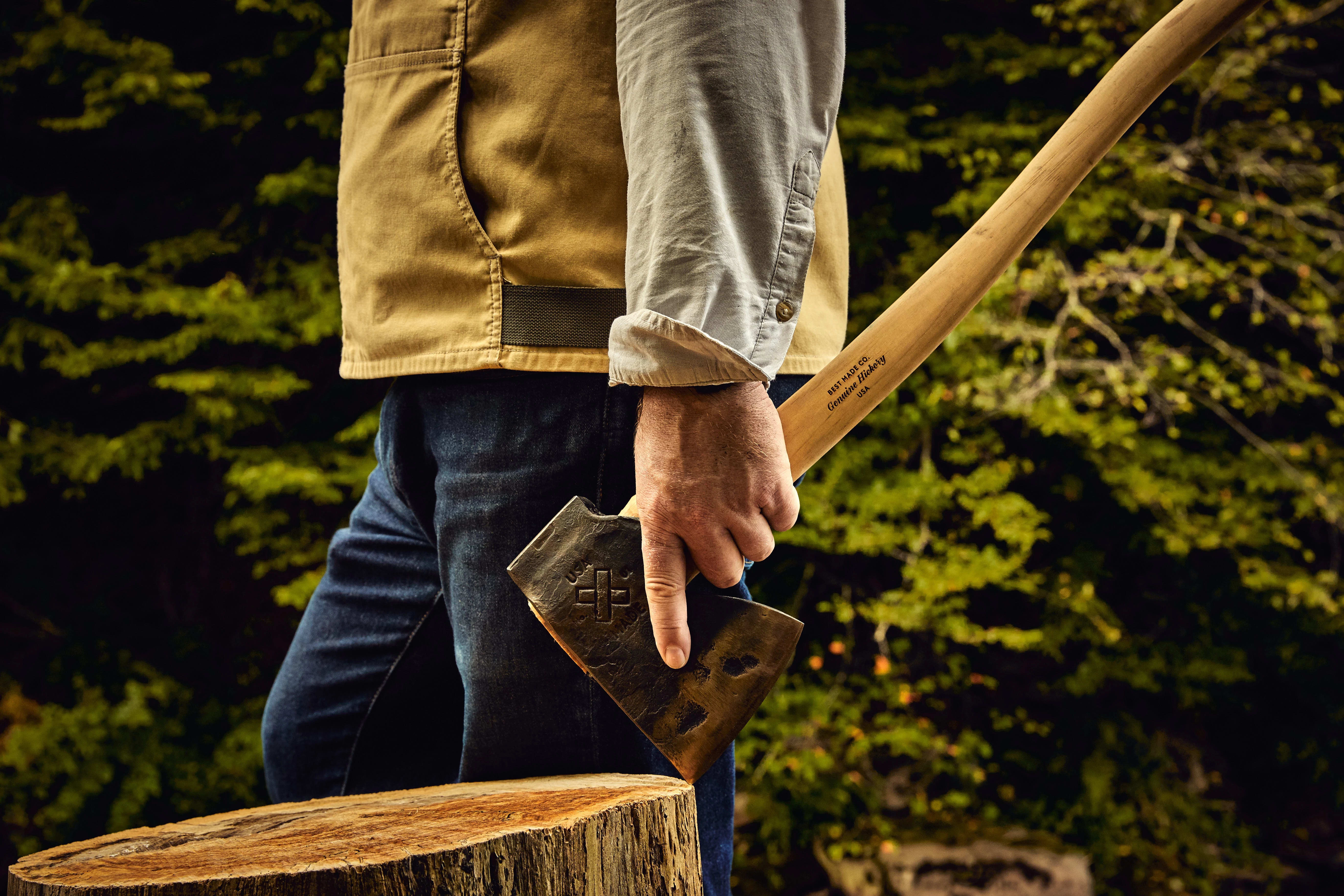 A person standing outdoors holds an axe by their side, with a tree stump in front and trees in the background.