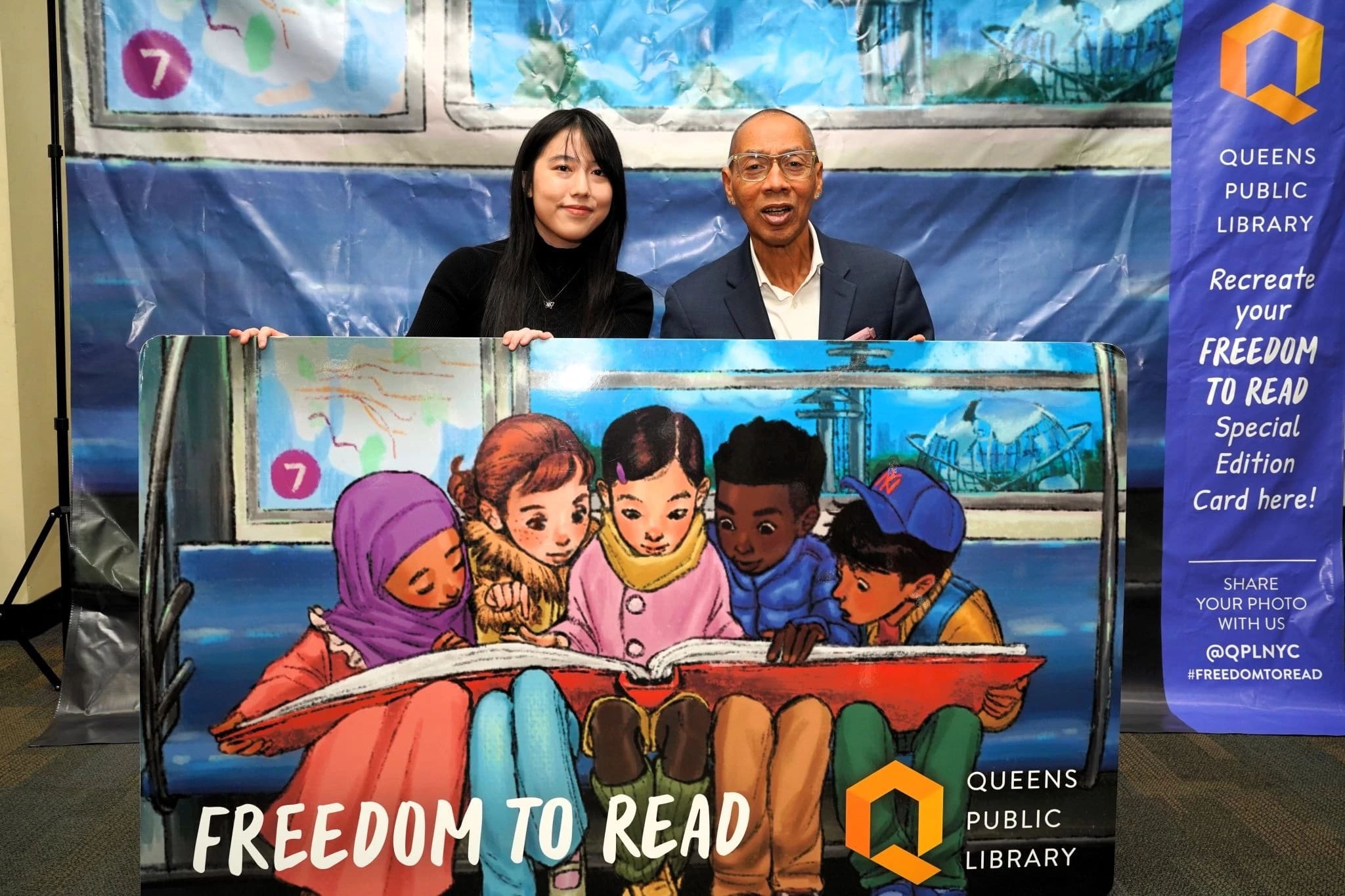 Sammi Wu sits besides a man in a suit holding a large version of a library card with an illustration of kids looking at a large red book.