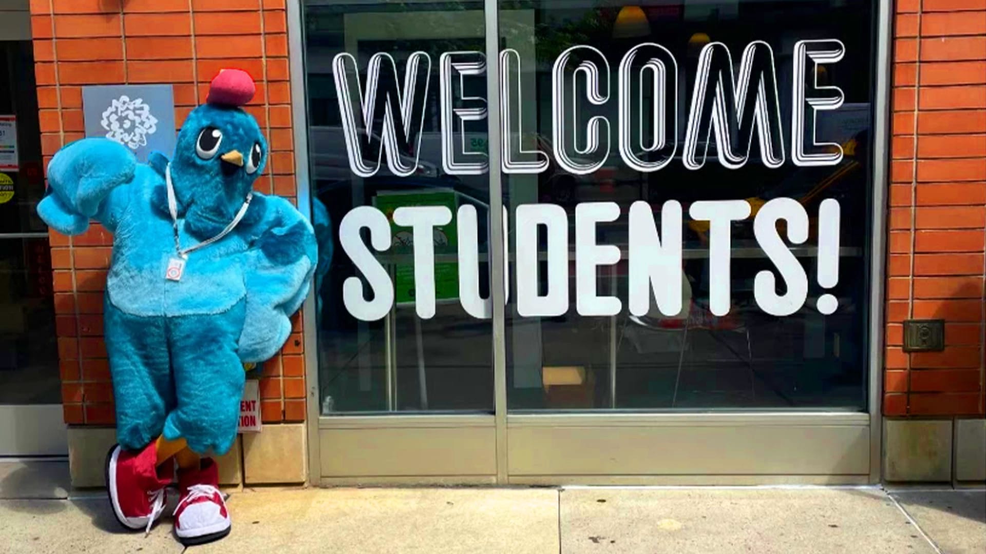 A bright blue pigeon mascot leans against a brick wall next to a window with "Welcome Students" on it in big letters.