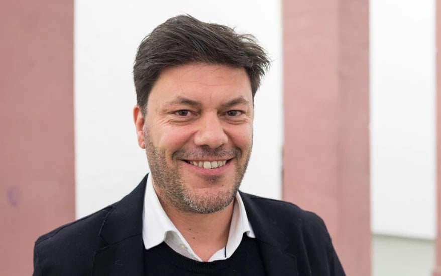 a smiling person with short brown hair and a black blazer on stands in front of a pink and white building exterior 