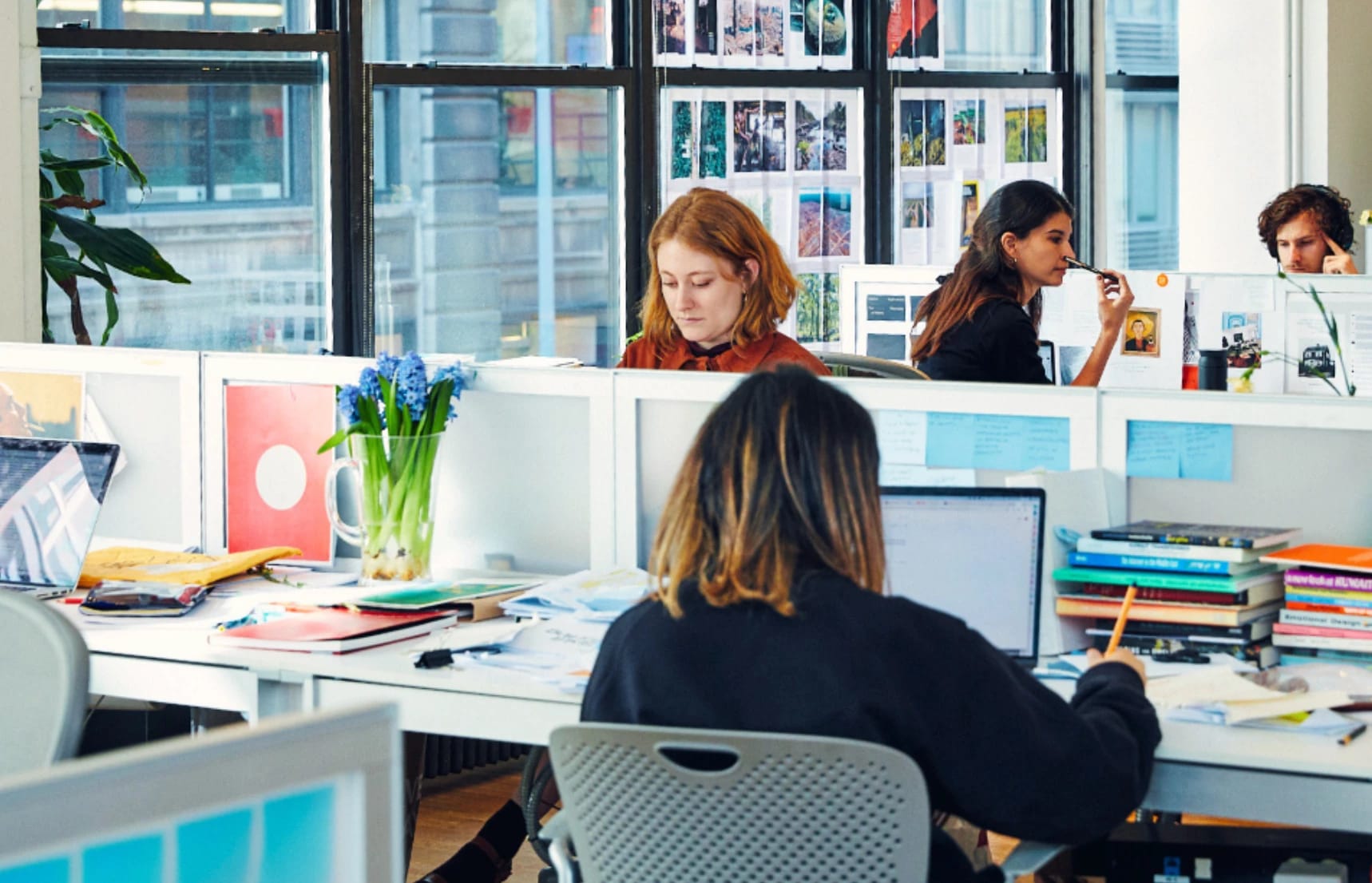 A person with orange hair sits at a row of desks working while other people in the room also work  
