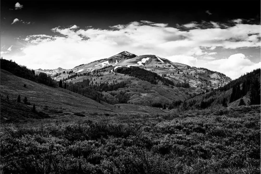 A black-and-white photograph of a mountainous landscape. A wooded valley is in the foreground and a snowy mountain is in the distance.