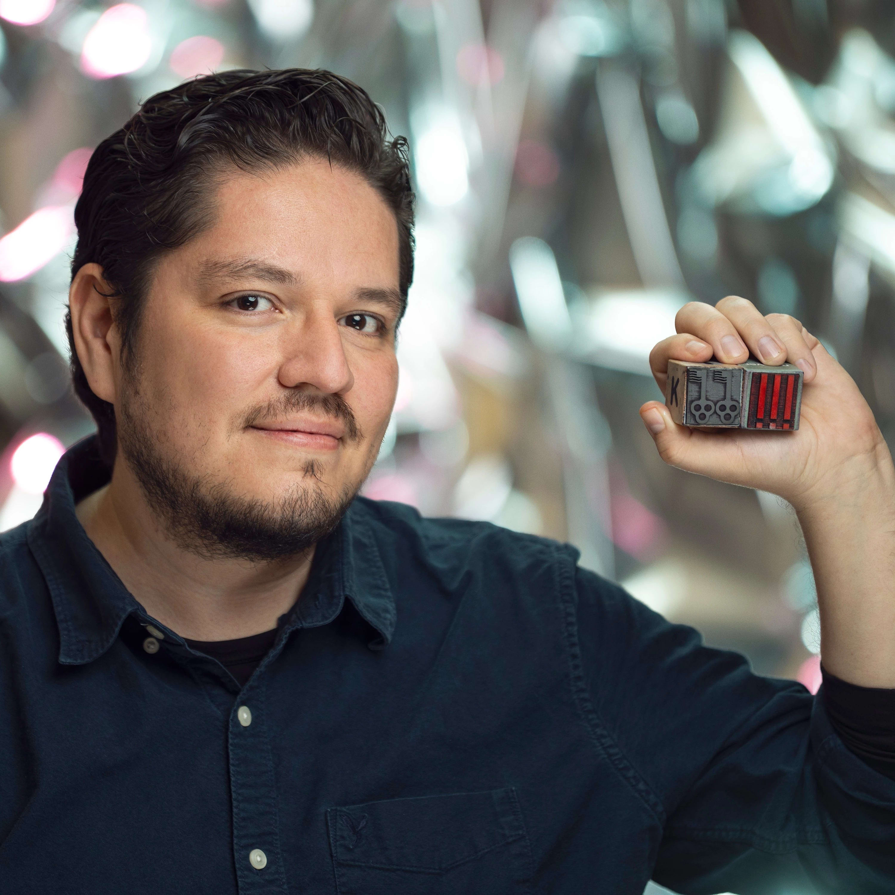 Photographic portrait of a person with dark hair and a deep blue shirt holding what appear to be oversized, illustrated die
